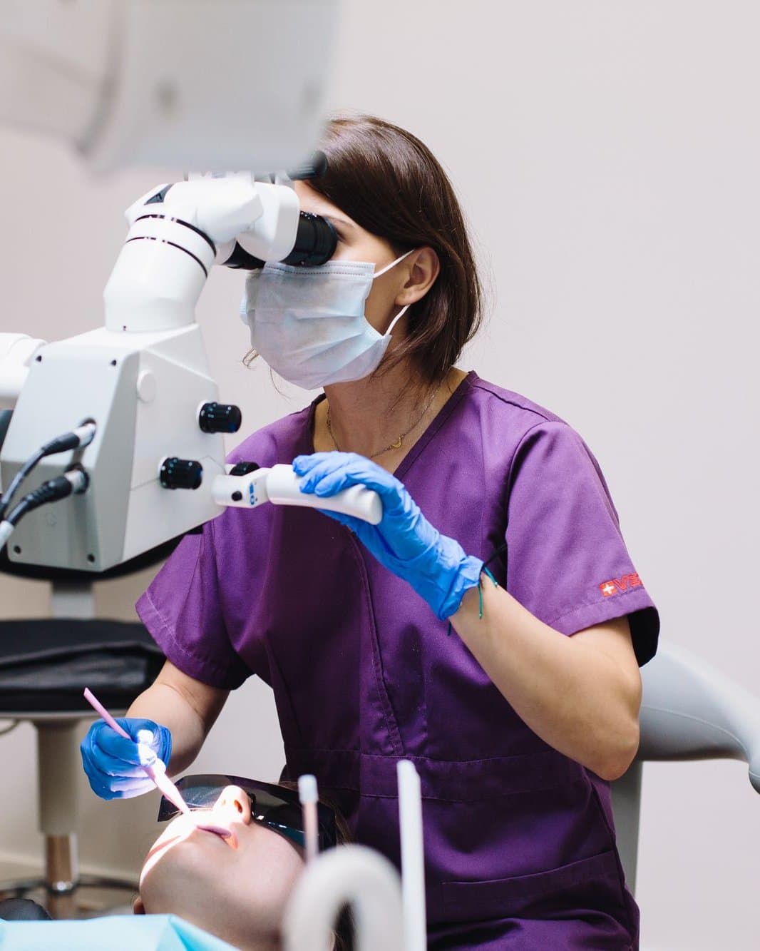 Dentista de uniforme roxo atendendo com o auxílio de um microscópio